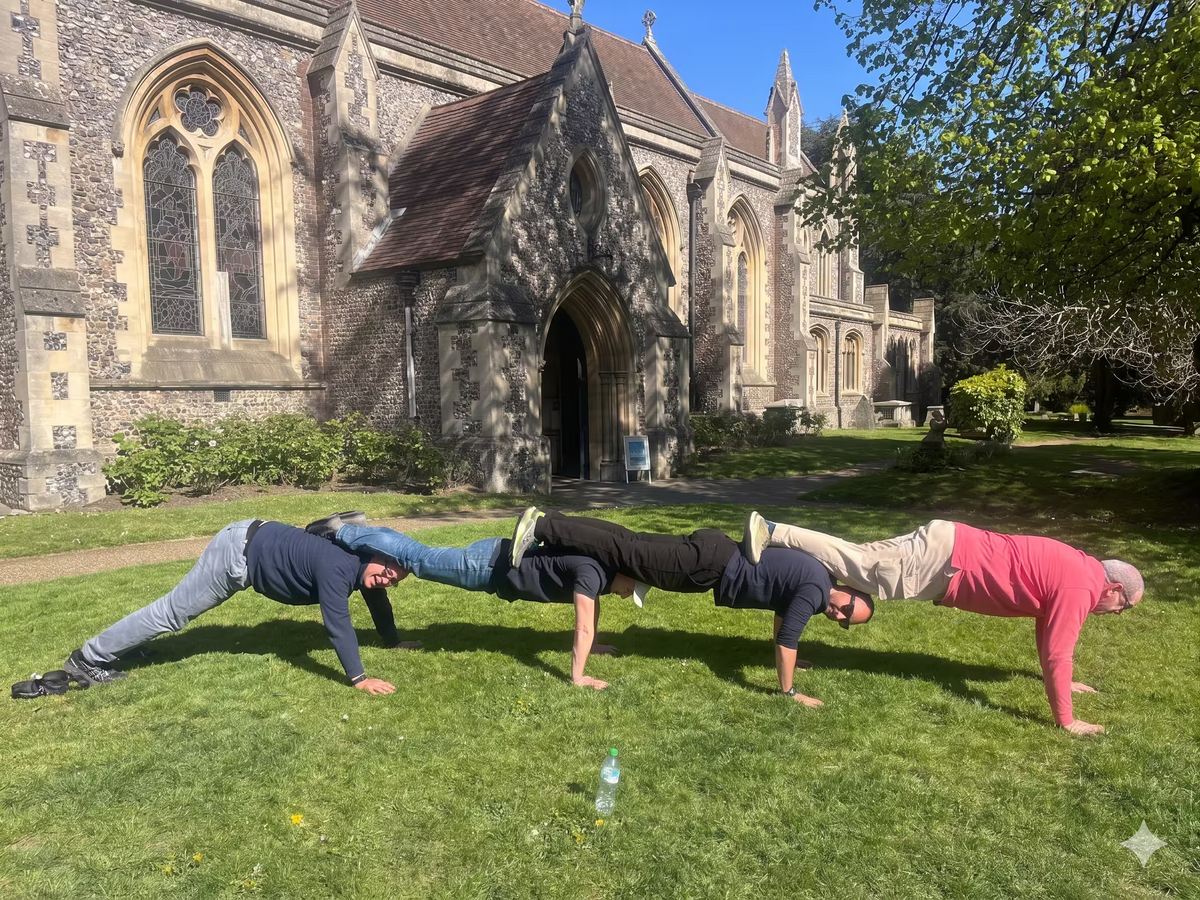 A team of people forming a connected "plank" line on the grass outside a church.