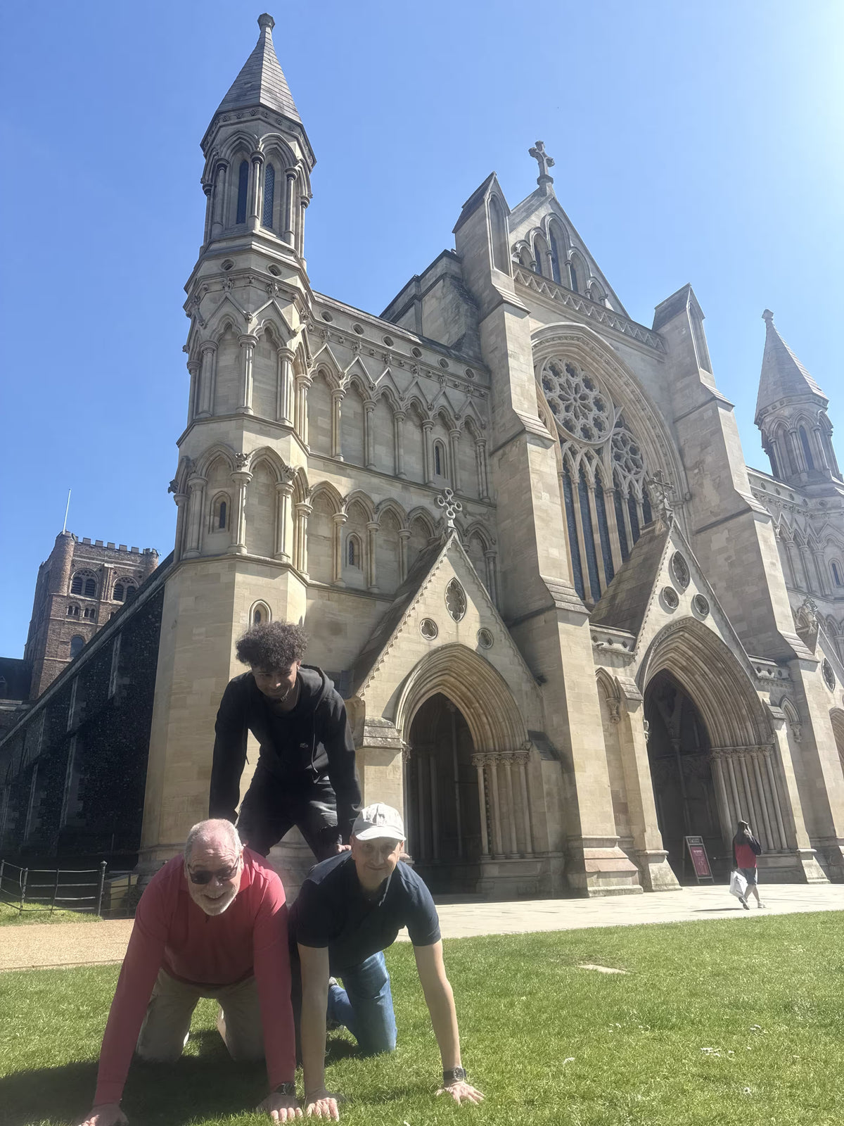 A small team of three people forming a human pyramid on a grassy lawn in front of a historic stone cathedral during a city scavenger hunt.