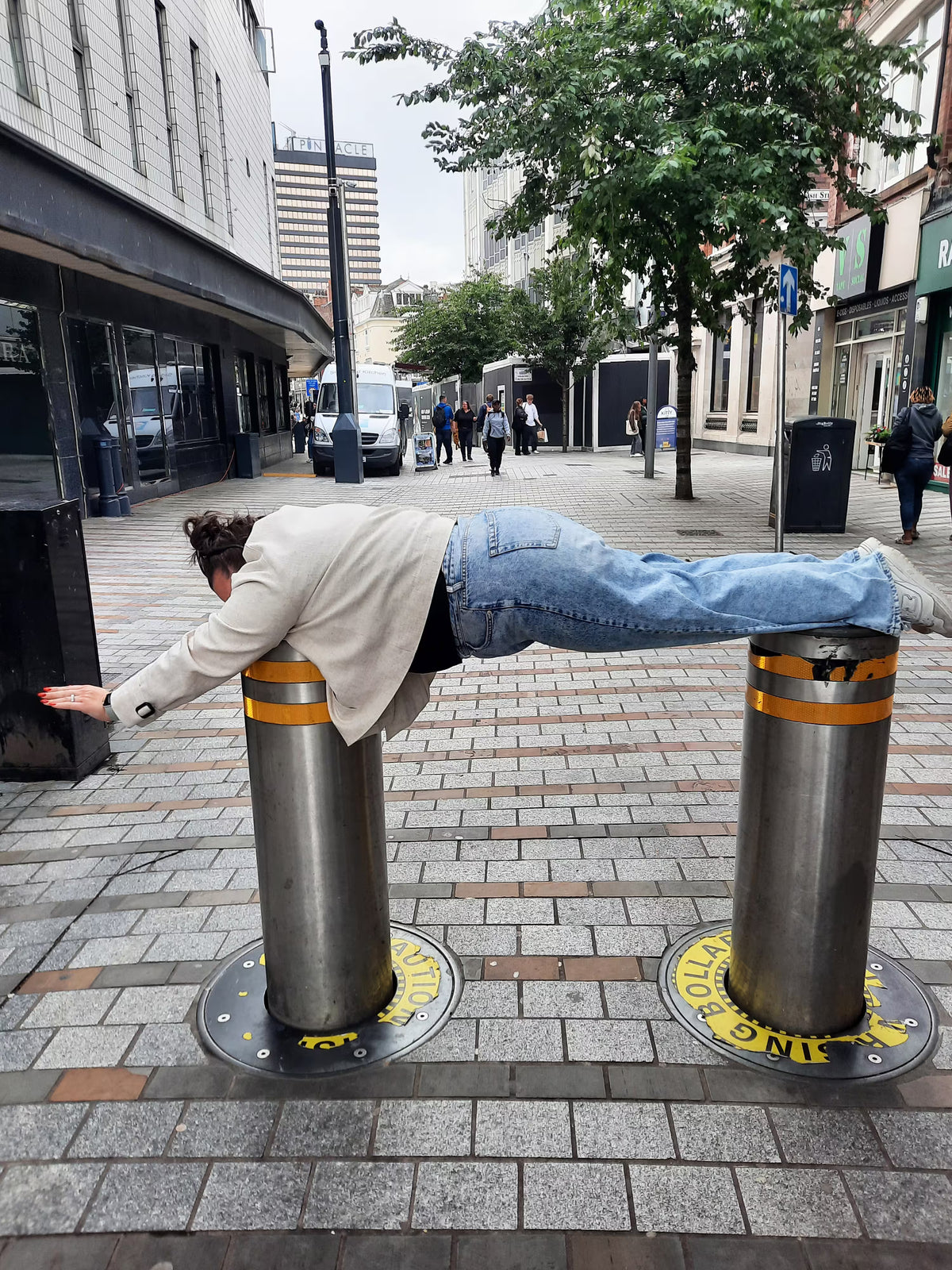 A person performing a "plank" across two metal street bollards in a city center.