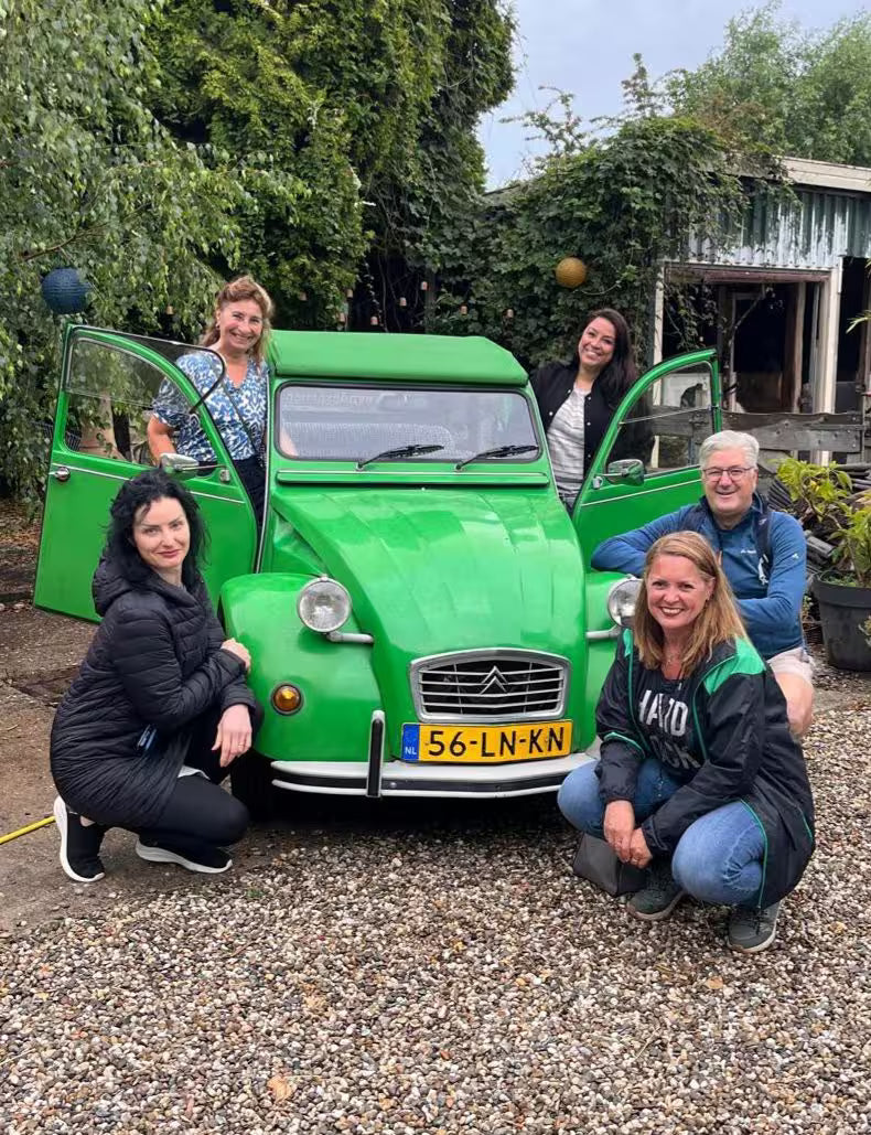 A group of colleagues posing around a small green vintage car during an outdoor event.